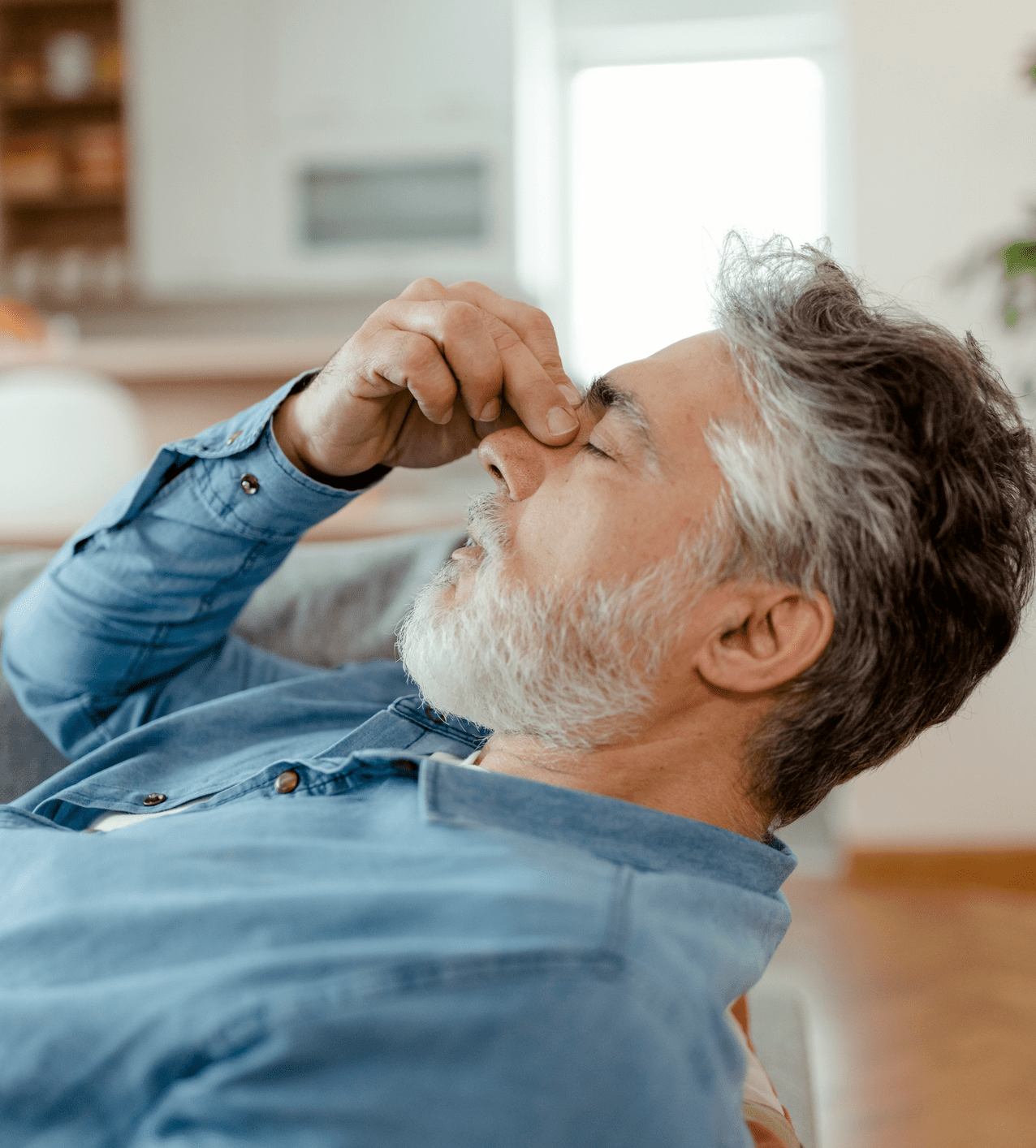 Older man with gray hair and beard holding his nose, appearing stressed or tired.